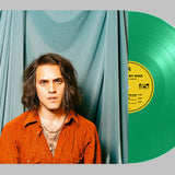 Person with long hair wearing an orange shirt in front of a green vinyl record with a yellow label.