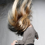 Person with long blonde hair blowing in the wind against a neutral background