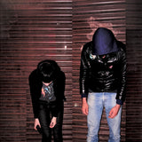 Two people in dark clothing against a corrugated metal wall.