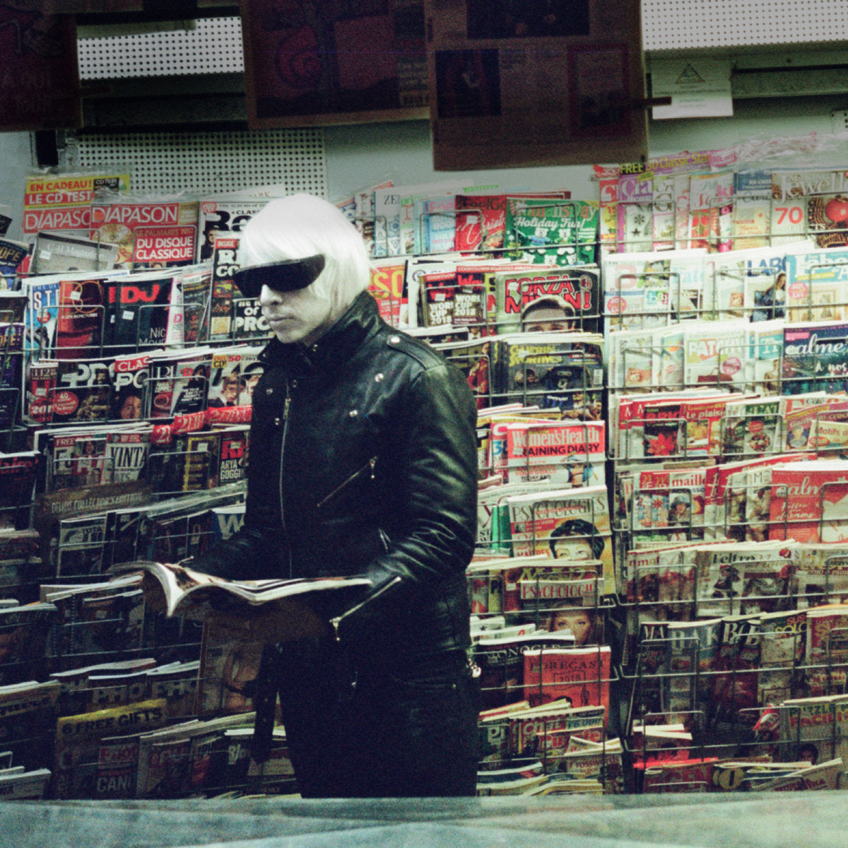 Album cover for Drab Majesty's "Modern Mirror" with a person in a black leather jacket and sunglasses standing in a magazine rack