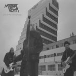 Album cover for Molchat Doma's 'Live At The Panorama Hotel' featuring the three musicians performing outdoors with a tall building in the background