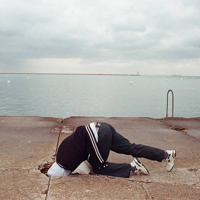 Soft Kill - Canary Yellow (Limited Edition) album cover with a person in tracksuit with their head in a cracked concrete surface by a body of water under an overcast sky.